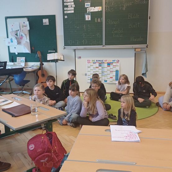 Ein Klassenzimmer mit Schülern, die auf dem Boden sitzen, einer hält ein Papier. Ein Tisch hat einen Rucksack und eine Gitarre ist platziert. Es gibt zwei Tafeln und ein Bulletin Board an der Wand.