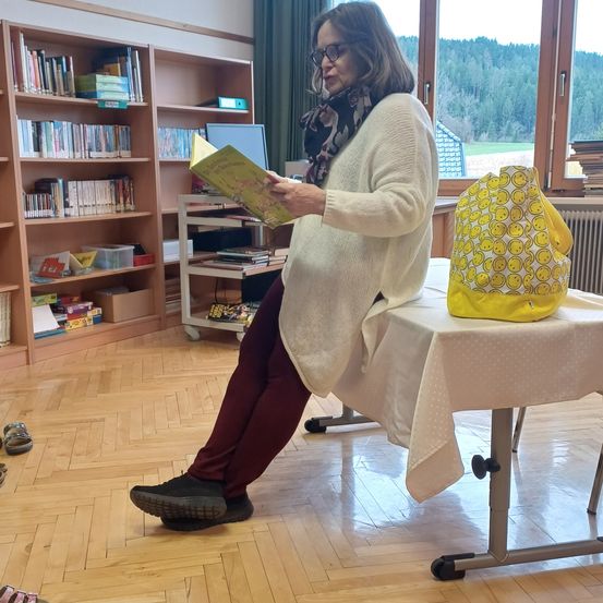Eine erwachsene Frau mit Brille und Schal sitzt in einem Klassenzimmer und liest ein Buch. Vor ihr steht ein Tisch mit einer gelben Tasche. Dahinter befinden sich Bücherregale, die mit Büchern und anderen Materialien gefüllt sind.