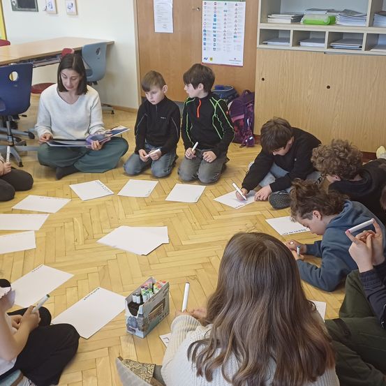 Ein Klassenzimmer mit Kindern, die in einem Kreis auf dem Boden sitzen und eine Zeichenaktivität durchführen. Papiere und Marker sind verteilt. Eine Frau sitzt in der Mitte mit einem Buch.