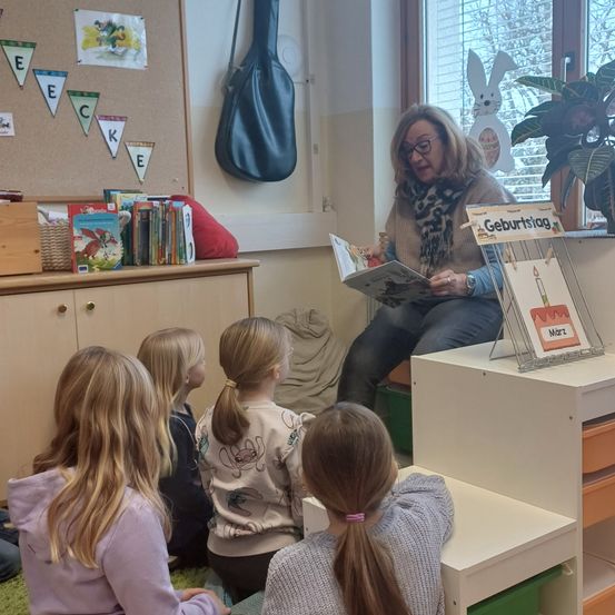 Eine Frau liest Kindern in einem Klassenzimmer ein Buch vor. Eine Pflanze und eine Gitarrentasche sind in der Nähe.