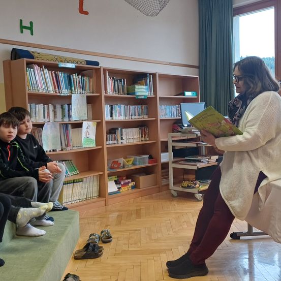Eine Frau liest drei Jungen in einer Bibliothek ein Buch vor. Die Kinder sitzen mit ausgezogenen Schuhen auf einer Bank. Die Frau steht und liest aus einem Buch vor.