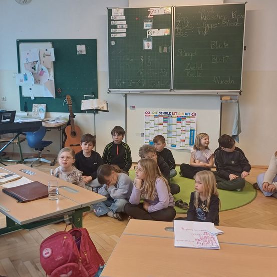 Ein Klassenzimmer mit Kindern auf dem Boden sitzend, einige mit gesenktem Kopf. Eine Lehrkraft unterrichtet. Eine Tafel mit deutschen Wörtern und Zahlen an der Wand.