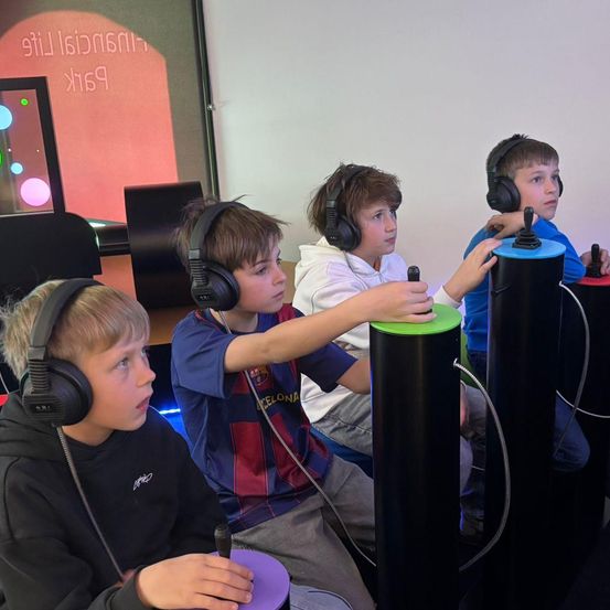 Vier Jungen spielen ein Videospiel, tragen Kopfhörer und halten Controller in den Händen. Sie sitzen um einen Tisch herum, mit Monitoren und einer Projektionsleinwand im Hintergrund.