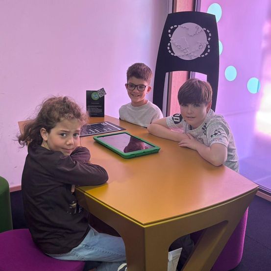 Drei Kinder sitzen um einen Tisch mit einem Tablet und lächeln in die Kamera. Hinter ihnen sorgt eine lila Wand mit bunten Lichtern für einen verspielten Effekt in der Szene.
