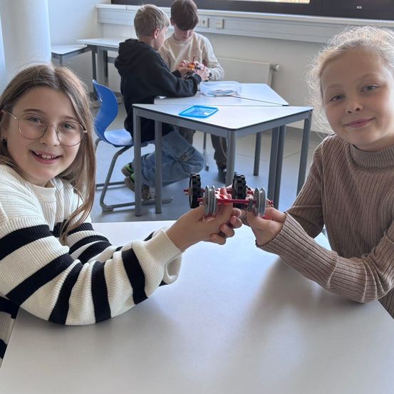 Zwei Mädchen halten ein rotes Spielzeugauto und lächeln. Dahinter sitzen zwei Jungen an Schreibtischen, einer arbeitet an einem Spielzeugauto. Sie sind in einem Klassenzimmer mit weißen Wänden und einem Fenster.
