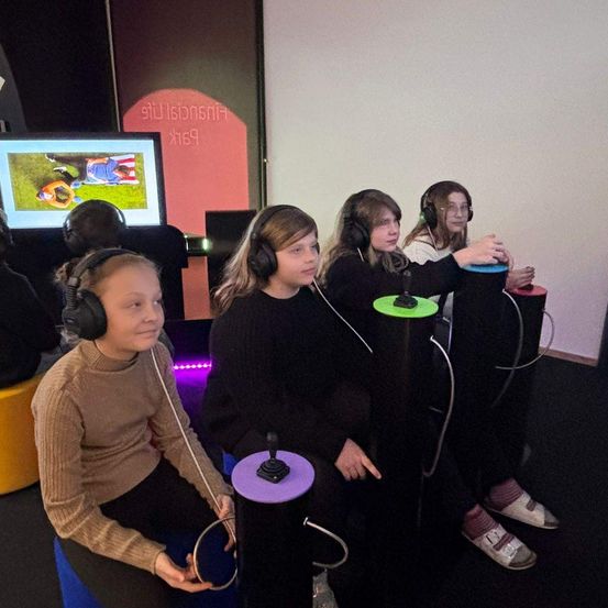 Vier Kinder sitzen und tragen Kopfhörer in einem Spielzimmer. Sie spielen ein Spiel auf einem großen Monitor.