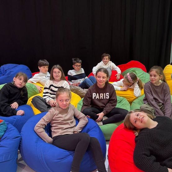 Eine Gruppe von Kindern, die auf bunten Beanbags mit schwarzem Hintergrund sitzen. Einige lächeln, andere sehen ernst aus. Ein Mädchen in einer schwarzen Paris-Sweatshirt.
