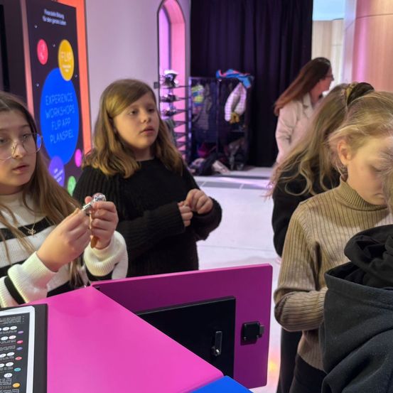 Drei junge Mädchen untersuchen kleine Objekte an einem rosa Kiosk in einem Geschäft. Ein Mädchen hält ein kleines Objekt und betrachtet es aufmerksam.