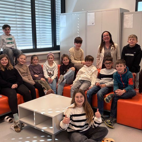 Eine Gruppe von Kindern und einer Frau sitzen auf orangefarbenen Sofas in einem Raum. Ein Mädchen gibt im Vordergrund einen Daumen hoch.