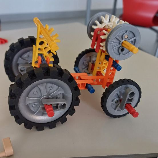 Ein farbenfrohes Spielzeugfahrzeug mit vier Rädern, roten und gelben Achsen und mehreren Zahnrädern steht auf einem weißen Tisch.