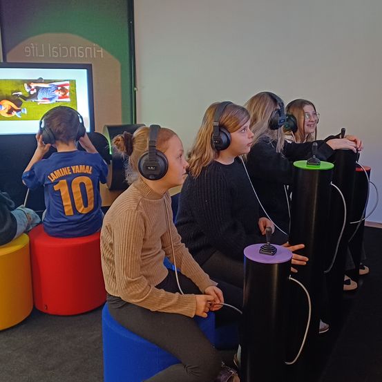 Fünf Kinder sitzen auf bunten Hockern, tragen Kopfhörer und spielen ein Videospiel. Der Bildschirm hinter ihnen zeigt eine Sportszene. Sie scheinen auf das Spiel konzentriert zu sein, mit Controllern in den Händen.