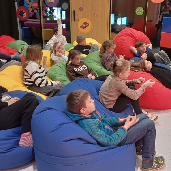 Kinder sitzen auf farbenfrohen Sitzsäcken in einem Spielzimmer, einige von ihnen unterhalten sich und andere sitzen ruhig da.