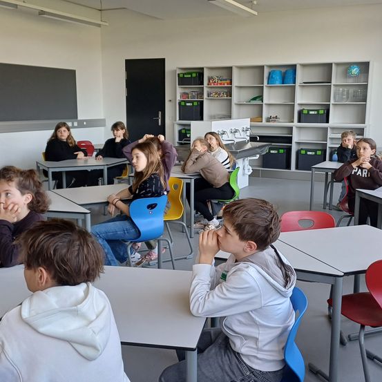 Ein Klassenzimmer mit Schülern, die an Tischen sitzen, einige mit den Händen vor dem Gesicht, weiße Wände, Regale und eine Tür im Hintergrund.