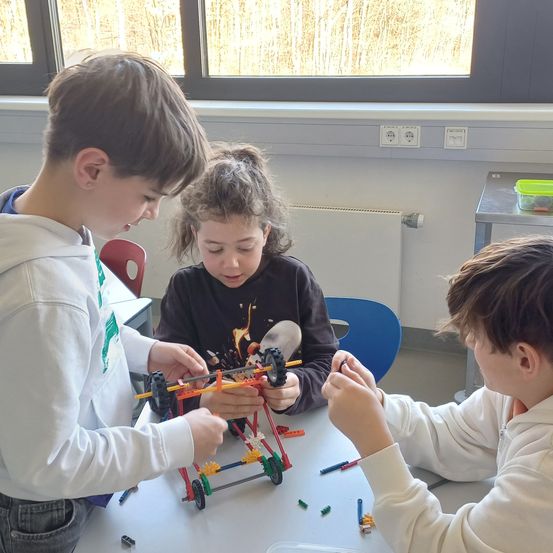 Drei Kinder sind drinnen und arbeiten an einem Lego-Fahrzeug. Sie sitzen an einem Tisch, wobei ein Kind eine grüne Lanyard trägt. Der Tisch ist mit Lego-Steinen und anderen Spielzeugen übersät.