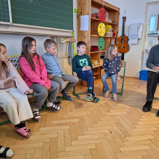 In einem Klassenzimmer sitzen mehrere Kinder auf Stühlen vor einem Erwachsenen, der ein Buch liest. Im Hintergrund befinden sich eine Tafel, ein Regal mit Musikinstrumenten und ein Papierspender.