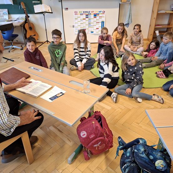 Eine Gruppe von Kindern sitzt in einem Kreis um einen Mann, der ein Buch liest. Sie befinden sich in einem Klassenzimmer mit Holzboden, einer Tafel und verschiedenen Schulmaterialien.