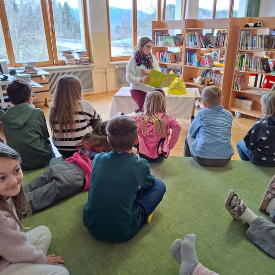 Eine Frau liest Kindern ein Buch vor in einer Bibliothek mit Bücherregalen und Fenstern, die einen Blick auf das Freie zeigen.