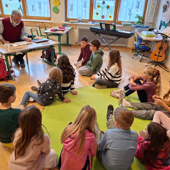 In einem Klassenzimmer sitzen eine Gruppe Kinder auf dem Boden um einen grünen Teppich herum, während ein Mann an einem Schreibtisch ein Buch liest. Im Hintergrund befinden sich Musikinstrumente, darunter eine Gitarre.