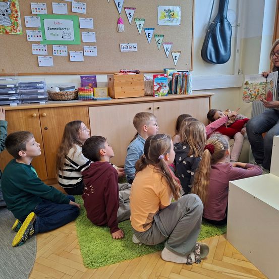 Ein Klassenzimmer, in dem eine Lehrerin ein Buch liest, während eine Gruppe von Kindern auf dem Boden sitzt. Im Hintergrund befindet sich eine Pinnwand mit bunten Papieren und einem Etui für eine Gitarre.
