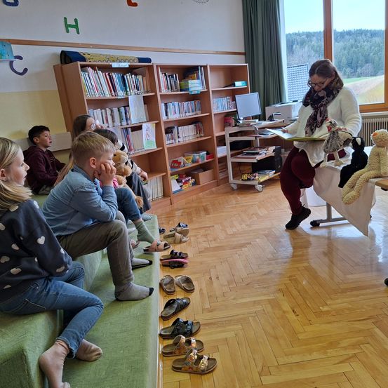 In einem Klassenzimmer sitzen Kinder barfuß auf Bänken und hören einer Frau zu, die ein Buch liest. Hinter ihnen stehen Regale mit Büchern. Sandale sind ordentlich auf dem Boden angeordnet.