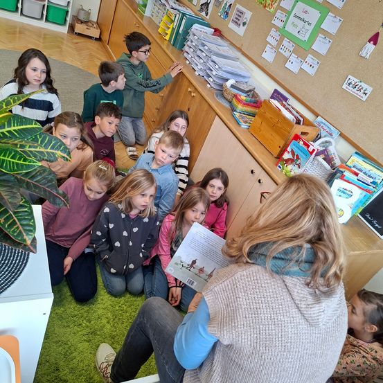 Eine Frau liest ein Buch vor einer Gruppe junger Kinder, die auf einem Teppichboden in einem Klassenzimmer sitzen.