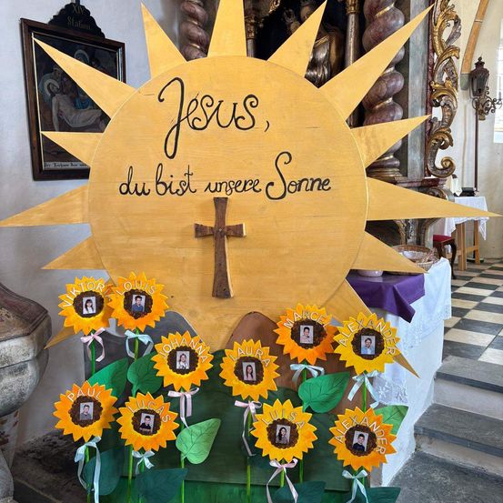 Eine gelbe Sonnenblumendekoration mit 'Jesus' darauf, umgeben von Fotos von Kindern. In der Mitte steht ein Kreuz. Links ist ein gerahmtes Bild von Jesus am Kreuz.