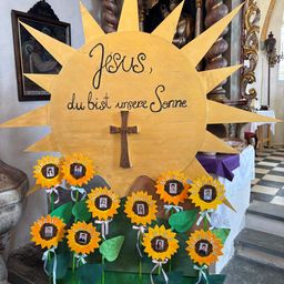 Eine gelbe Sonnenblumendekoration mit 'Jesus' darauf, umgeben von Fotos von Kindern. In der Mitte steht ein Kreuz. Links ist ein gerahmtes Bild von Jesus am Kreuz.