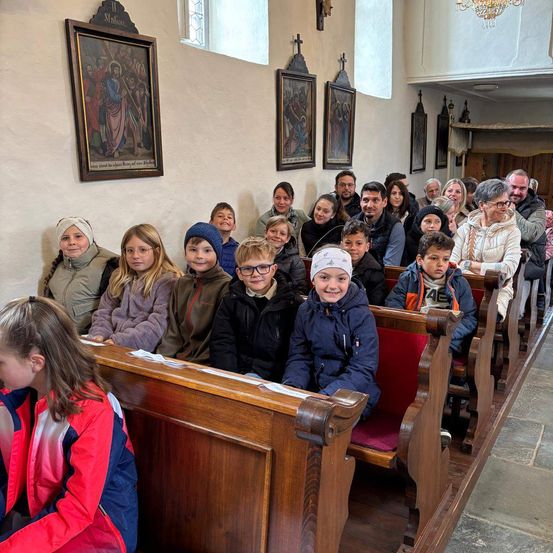 Eine Gruppe von Kindern und Erwachsenen sitzt in Kirchenbänken in einer Kapelle mit religiösen Ikonen und einer Kronleuchter.