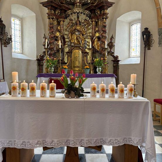 Ein weißer Altartisch, geschmückt mit Kerzen und Blumen, steht vor einem goldenen Altar. Die Kerzen sind mit Namen beschriftet, und eine Statue befindet sich hinter dem Altar. Die Umgebung ist in einer Kirche mit weißen Wänden und Buntglasfenstern.
