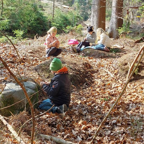 Vier Kinder sitzen auf einem Felsvorsprung im Wald. Drei von ihnen sind mit einer Aktivität beschäftigt. Sie sind von gefallenen Blättern und Zweigen umgeben. Ein Weg und ein Baumstumpf sind in der Ferne sichtbar.