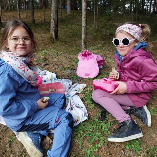 Zwei kleine Mädchen sitzen auf dem Boden in einem Wald und essen von ihren Lunchboxen. Ein Mädchen trägt eine Brille, einen Schal und eine Jacke. Das andere Mädchen hat eine Stirnband, eine Sonnenbrille und eine rosa Jacke.