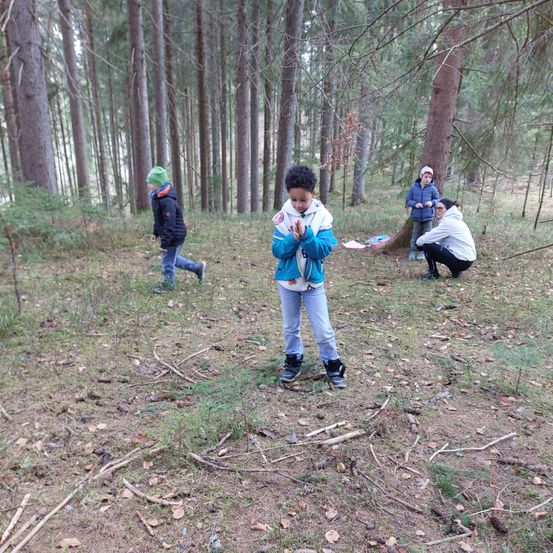 Vier Kinder in einem Wald. Ein Junge vorn betet, während andere gehen. Bäume umgeben das Gebiet.
