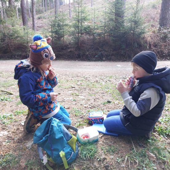 Zwei Kinder sitzen im Wald auf dem Boden, essen und trinken. Eines hat einen Rentierhut. Sie sind von Natur umgeben.