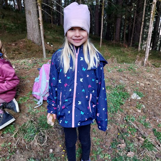 Ein junges Mädchen in einer blauen Jacke mit Schmetterlingsmuster und einer lila Mütze steht lächelnd im Wald.