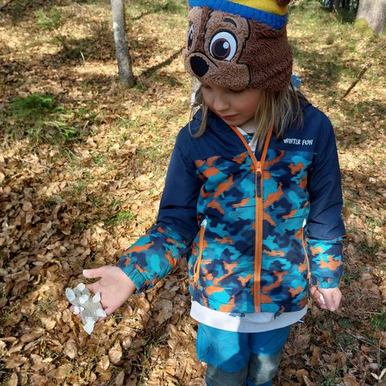 Ein Kind in einer Camouflage-Jacke und einem Bärenhut hält einen Stein in der Hand, während es auf einem Waldboden steht, der mit trockenen Blättern bedeckt ist.