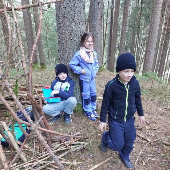 Drei Kinder im Wald, eines hält eine Kiste, ein anderes steht neben einem Baum und ein drittes läuft.