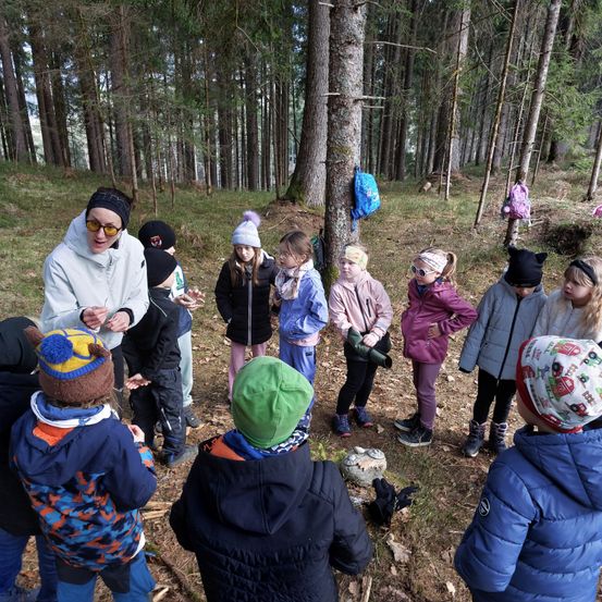 Eine Gruppe von Kindern und eine erwachsene Frau stehen in einem Wald. Sie tragen Winterkleidung. Die Frau erklärt den Kindern etwas.
