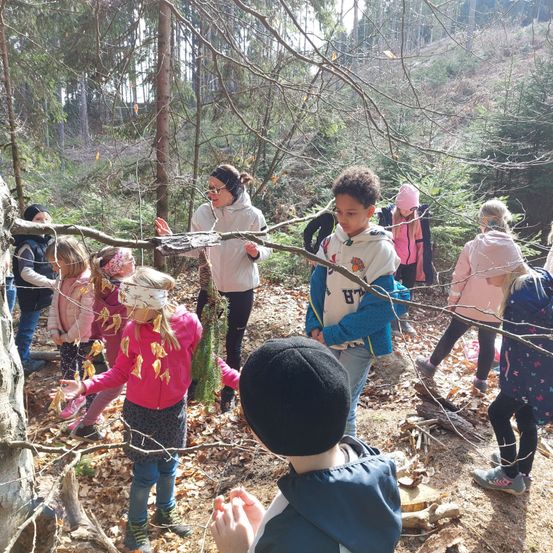 Eine Gruppe von Kindern und einem Erwachsenen ist in einem Wald und sammelt Blätter und Zweige, möglicherweise auf einem Naturspaziergang.