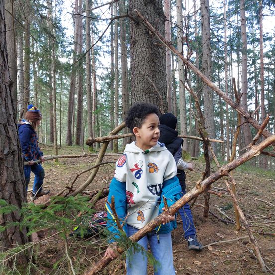 Kinder spielen im Wald und halten Stöcke und Äste. Ein Kind steht im Vordergrund und lächelt, während andere dahinter stehen. Sie scheinen etwas zu bauen.
