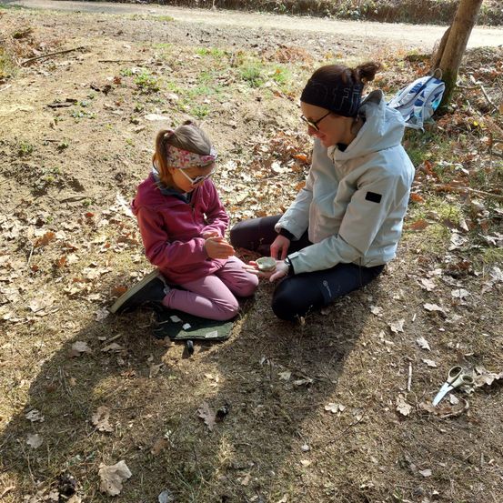 Eine Frau und ein Kind sitzen auf dem Boden und untersuchen etwas in ihren Händen. Sie sind von gefallenen Blättern umgeben und haben einen Rucksack in der Nähe.