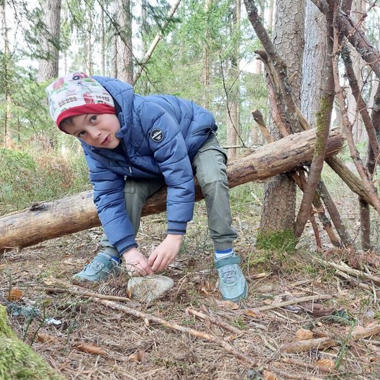 Ein junger Junge in einer blauen Jacke sitzt auf einem Baumstamm in einem Wald und baut möglicherweise ein Schutzdach aus Zweigen.
