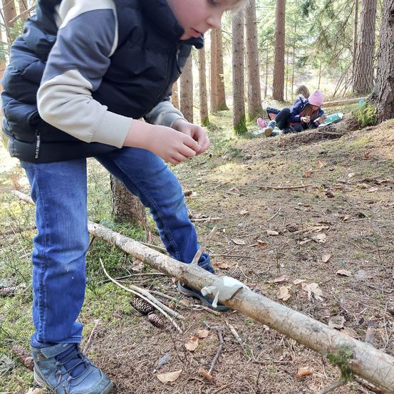 Ein Kind steht in einem Wald und hält einen Baumstamm. Hinter ihm sitzt ein Mädchen auf dem Boden. Der Wald ist von hohen Bäumen umgeben.