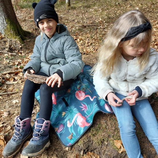 Zwei junge Mädchen sitzen auf einer Decke in einem bewaldeten Gebiet, eine untersucht einen Stock, die andere hält einen Stein. Sie tragen Winterkleidung und sind von gefallenen Blättern umgeben.