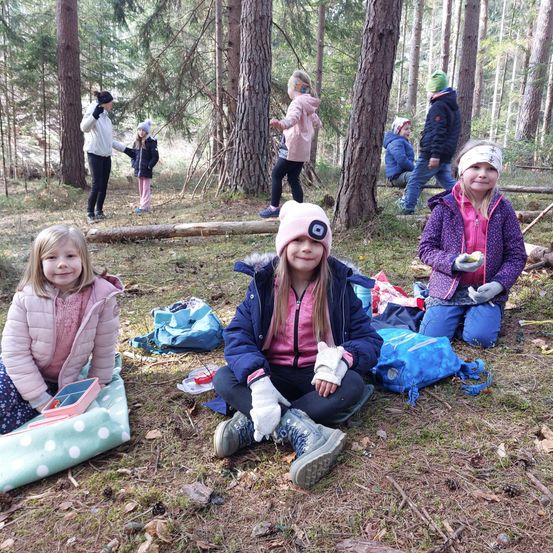 Kinder in Winterkleidung sitzen im Wald. Eines hält eine rosa Schachtel, ein anderes trägt einen rosa Hut. Zwei weitere laufen im Hintergrund, und einer hält einen Baumstamm.
