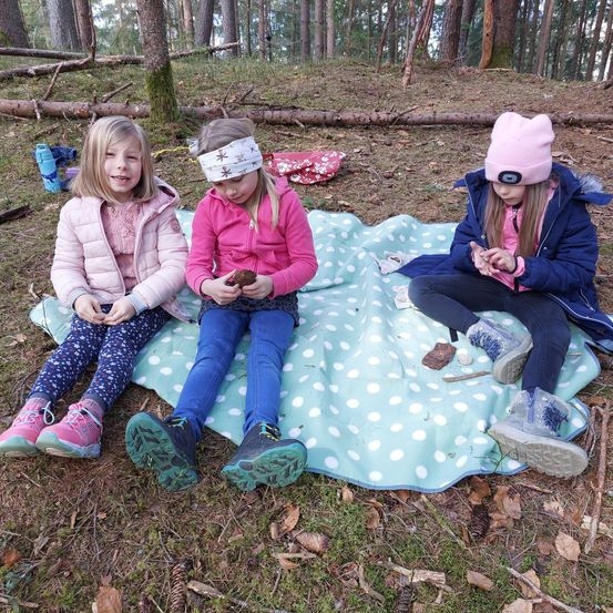 Drei junge Mädchen sitzen auf einer Decke im Wald. Eine hält ein kleines Objekt. Sie tragen Winterkleidung und haben einen fröhlichen Ausdruck.