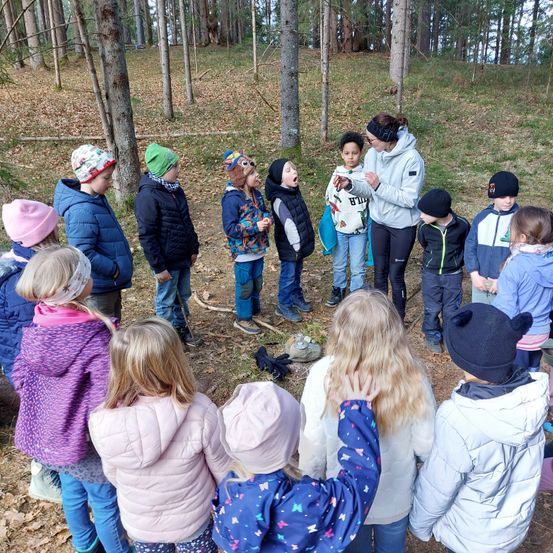 Eine Gruppe von Kindern in warmer Kleidung steht im Kreis im Wald und hört einem Erwachsenen zu, der ein Objekt hält. Die Kinder tragen verschiedene Mützen, und der Boden ist mit Blättern bedeckt.