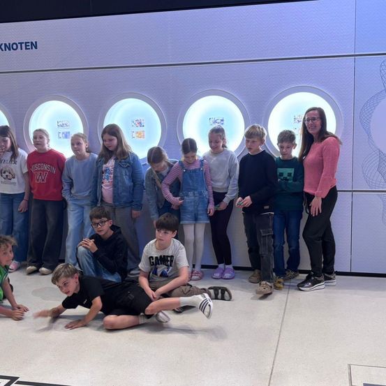 Eine Gruppe von Kindern und einer erwachsenen Frau posiert vor einer Wand mit kreisförmigen Lichtern. Die Kinder tragen Freizeitkleidung und Sneakers, während die Frau in einem rosa Top ist.