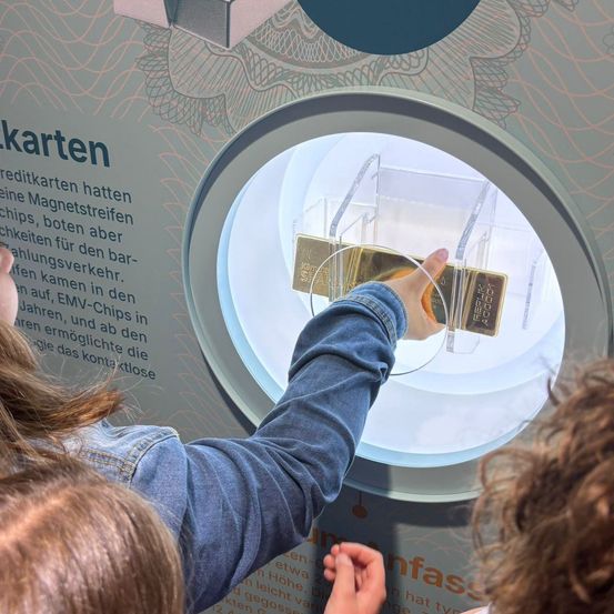 Zwei Personen untersuchen eine goldene Barren in einem durchsichtigen Halter durch ein kreisförmiges Fenster an einer Wand. Die Wand hat Text auf Deutsch, der möglicherweise die Geschichte von Kreditkarten beschreibt.