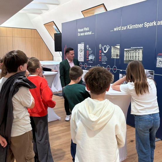 Eine Gruppe von Kindern und Erwachsenen versammelt sich in einem Raum und studiert eine Ausstellungstafel. Ein Mann steht vorne und erklärt möglicherweise die Ausstellung. Der Raum hat Holzböden und ein modernes Innendesign.