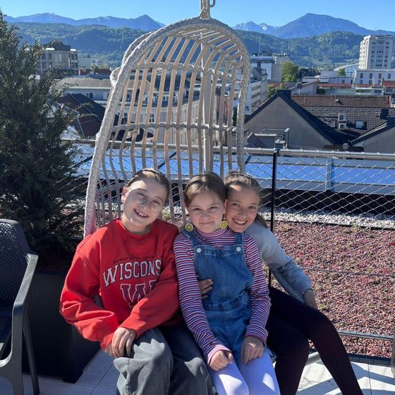 Drei Kinder sitzen auf einer Dachterrasse auf einem Stuhl und lächeln, mit einer Stadt und Bergen im Hintergrund.